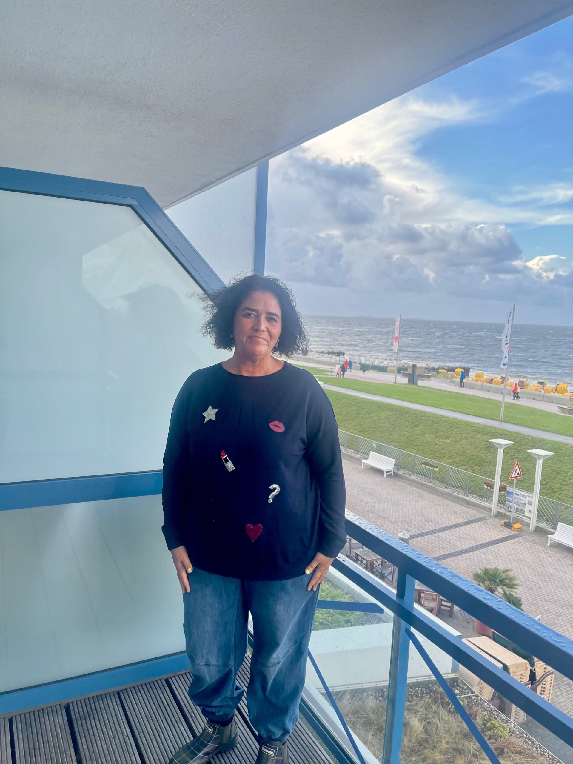 Nicole steht auf einem Balkon mit Blick auf Strandpromenade und Meer