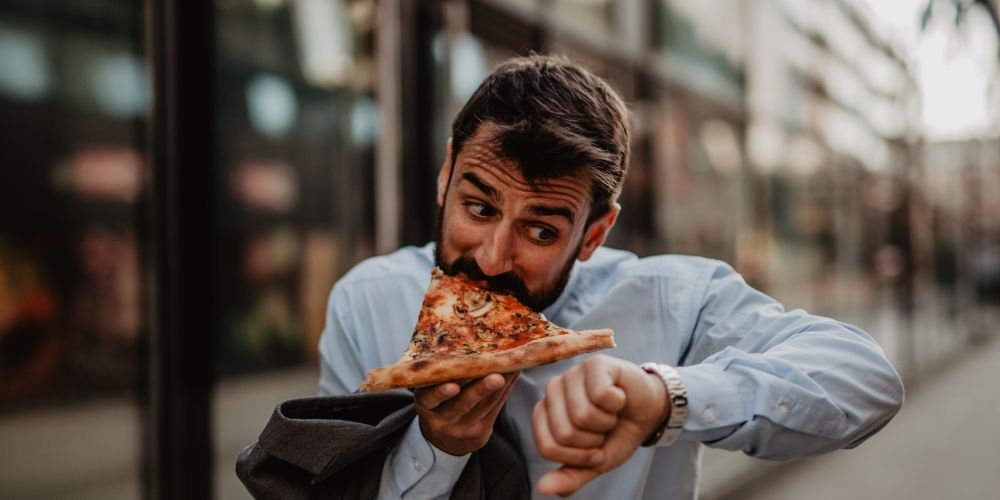 Mann isst hastig ein Stück Pizza und schaut dabei auf seine Uhr