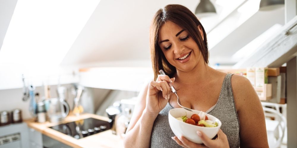 Frau isst lächelnd eine Schüssel Salat in ihrer Küche